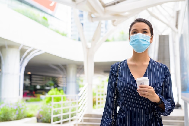 Giovane imprenditrice asiatica con maschera che beve caffè in movimento come la nuova normalità durante il covid-19