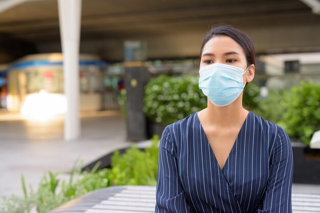 Giovane imprenditrice asiatica con maschera per la protezione dallo scoppio del virus corona pensando e seduto in città