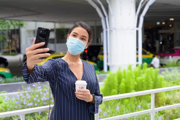 Giovane imprenditrice asiatica con maschera prendendo selfie mentre beve un caffè in movimento come la nuova normalità in città all'aperto