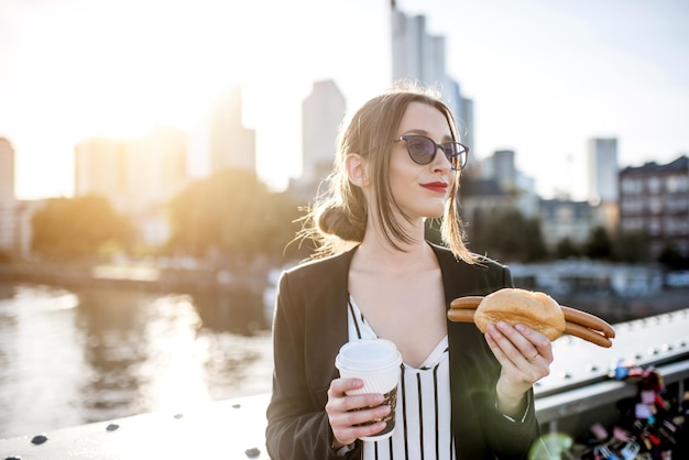 Giovane imprenditrice avente uno spuntino con salsiccia tradizionale Franfurt e caffè sul ponte con grattacieli sullo sfondo a Francoforte durante il tramonto