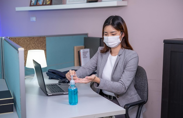 Giovane imprenditrice che indossa una maschera facciale e usa il gel per le mani disinfettante mentre lavora al computer