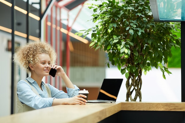 Giovane imprenditrice con capelli ricci seduto al suo posto di lavoro davanti al computer portatile e parlando al telefono cellulare