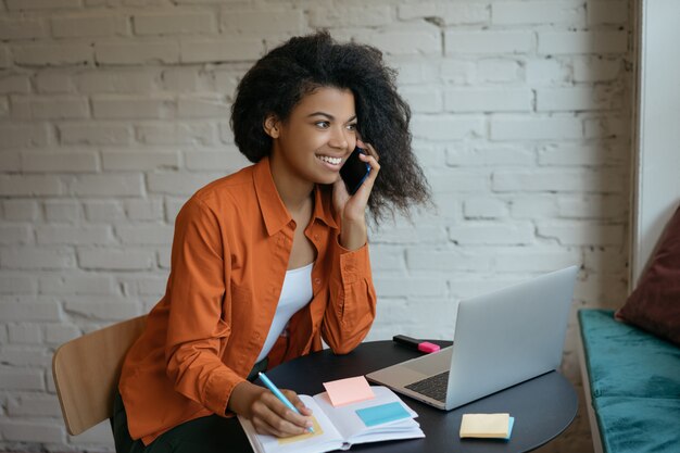 Giovane imprenditrice con laptop, parlando sul cellulare, prendere appunti, pianificare l'avvio