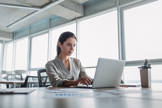 Giovane imprenditrice dai capelli scuri che indossa una camicia a righe è seduta nel suo ufficio e sta esaminando il computer portatile. Il suo taccuino e la matita sono distesi sulla scrivania. Ufficio moderno luminoso.