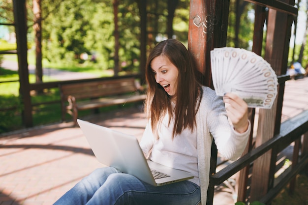 Giovane imprenditrice di successo che tiene un pacco di dollari, denaro contante. Donna che lavora al computer pc portatile moderno nel parco cittadino in strada all'aperto sulla natura. Ufficio Mobile. Concetto di business freelance.