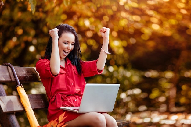 Giovane imprenditrice euforica che celebra il successo aziendale leggendo buone notizie sul laptop nel parco.