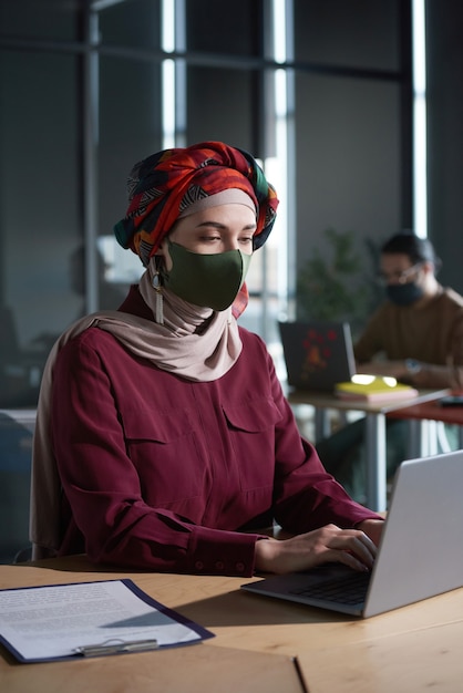 Giovane imprenditrice in hijab e maschera protettiva che utilizza laptop per il lavoro online durante la pandemia