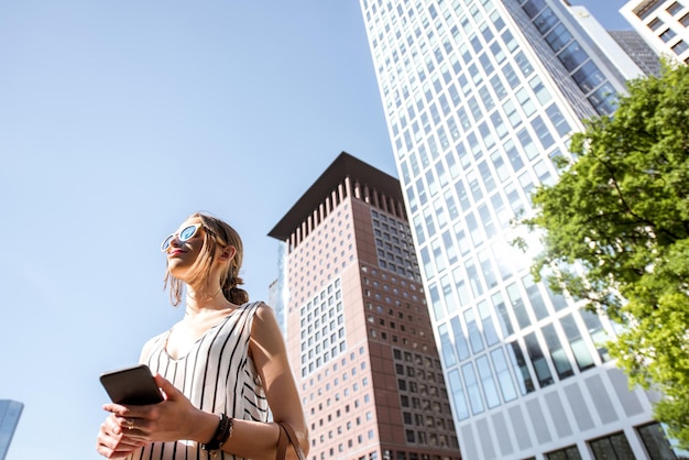 Giovane imprenditrice in piedi con lo smartphone all'aperto presso il moderno quartiere della città di Francoforte. Vista dal basso con grattacieli sullo sfondo