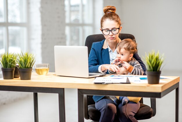 Giovane imprenditrice multitasking vestita con la tuta che lavora con laptop e documenti seduta con il figlioletto in ufficio