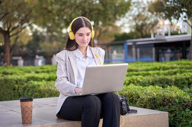 Giovane imprenditrice seduta sulla panchina guardando il suo computer portatile Bella donna che indossa abbigliamento formale utilizzando le cuffie