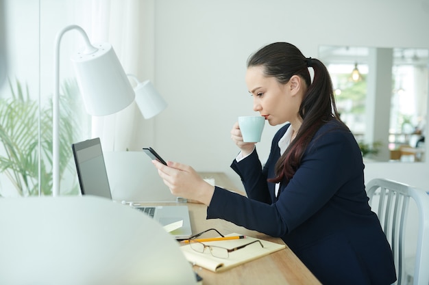 Giovane imprenditrice seria che beve caffè e legge un messaggio di testo da un collega o un partner commerciale