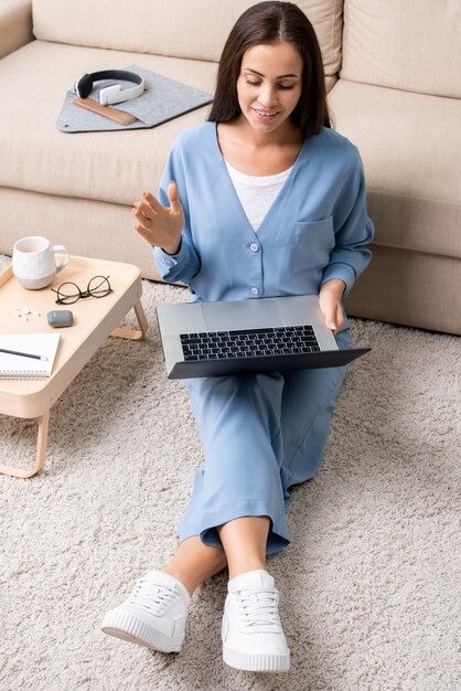 Giovane imprenditrice sorridente in pigiama blu agitando la mano durante la comunicazione online con i colleghi mentre è seduta sul pavimento vicino al divano