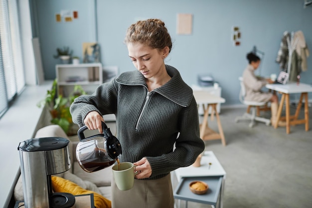 Giovane imprenditrice versando caffè caldo in tazza durante la pausa caffè in ufficio