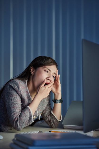 Giovane imprenditrice vietnamita che sbadiglia durante la lettura del documento di posta elettronica sul monitor del computer, sta lavorando in ufficio buio