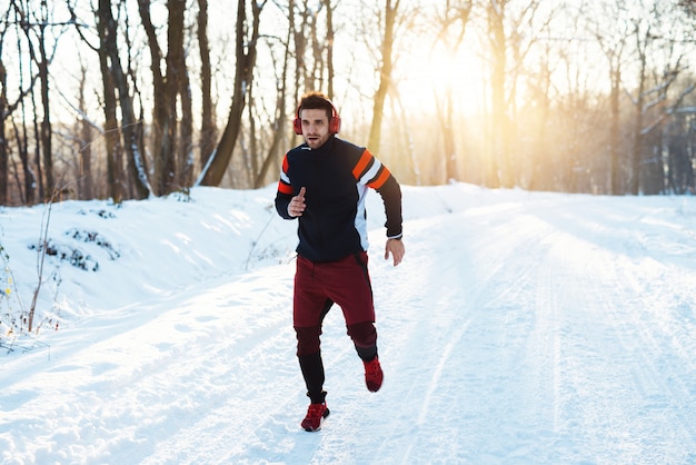 Giovane in buona salute in abiti sportivi con le cuffie che corre sulla strada innevata di inverno di mattina.