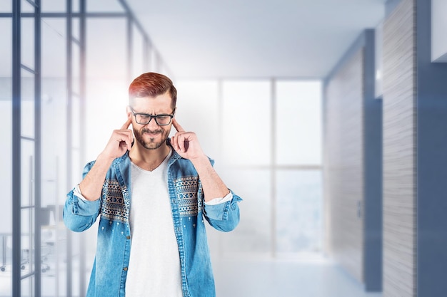 Giovane in camicia di jeans e occhiali che coprono le orecchie e in piedi con gli occhi chiusi nella hall dell'ufficio offuscata. Concetto di stress. Immagine tonica