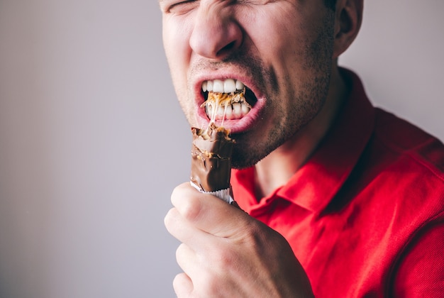 Giovane in camicia rossa isolata sopra la parete. Primo piano della bocca aperta per un'altra porzione di barretta di cioccolato.