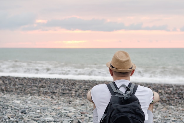 Giovane in cappello e con lo zaino che si siede sulla spiaggia. Mare e cielo al tramonto. Vista posteriore