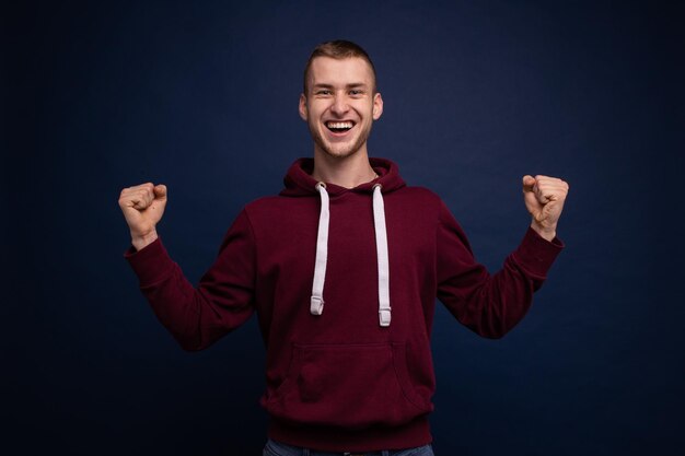 Giovane in felpa con cappuccio rossa e jeans in posa su sfondo blu sorridente che mostra la vittoria