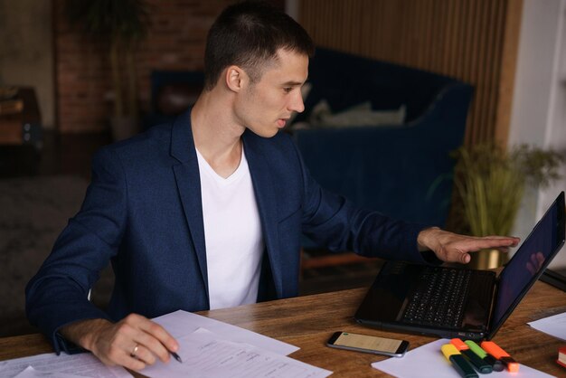 Giovane in giacca che lavora da casa facendo scartoffie utilizzando il computer portatile e tenendo