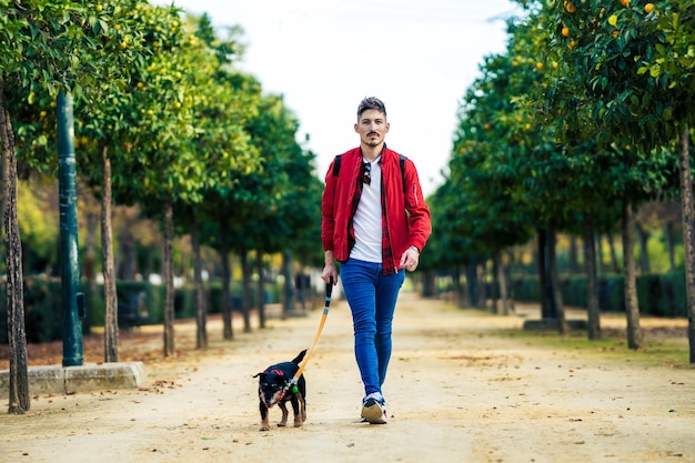 Giovane in giacca rossa che porta a spasso il cane attraverso un sentiero sabbioso di un parco circondato da alberi