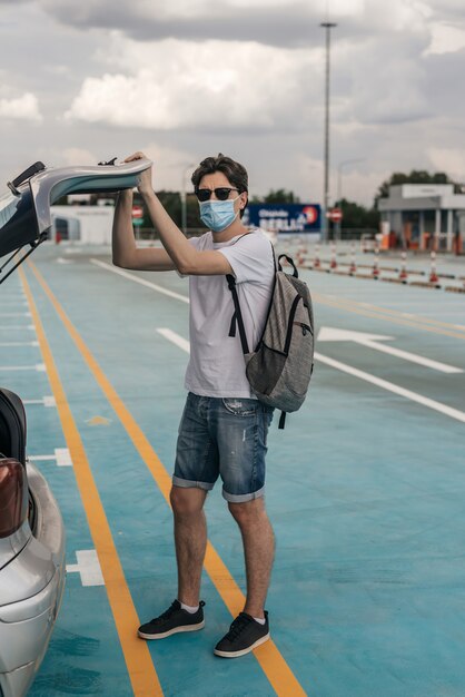 Giovane in maglietta bianca, pantaloncini di jeans, occhiali da sole e maschera chirurgica con zaino sulla schiena. Attraverso la vista auto