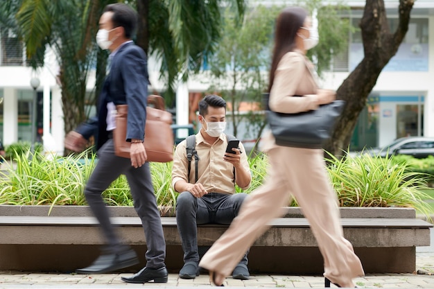 Giovane in maschera medica seduto su una panchina quando le persone che passano in movimento sfocato