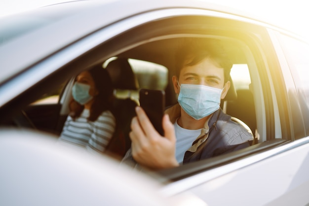 Giovane in maschera medica sterile protettiva che utilizza il telefono alla guida dell'auto.