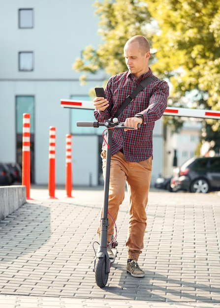 Giovane in pantaloni casual e camicia in piedi accanto al suo scooter elettrico, controllando il telefono cellulare