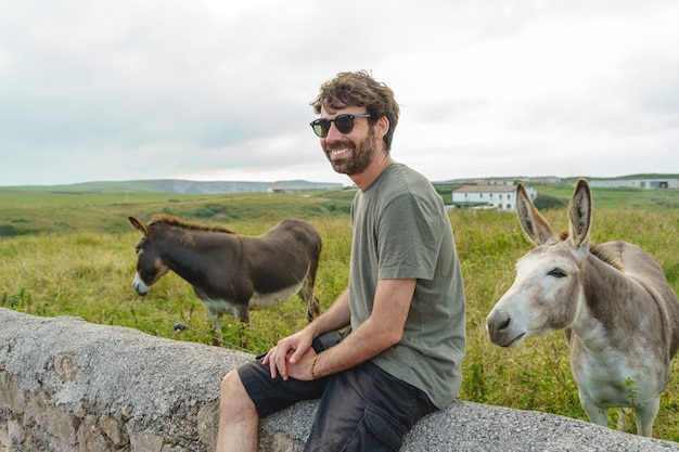 Giovane in un prato con gli asini. Vista orizzontale dell'uomo in vacanza in campagna con animali.