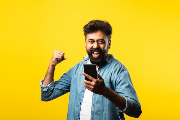 Giovane indiano barbuto che usa lo smartphone, sorride mentre chiama o chiacchiera con un amico, in piedi sul giallo