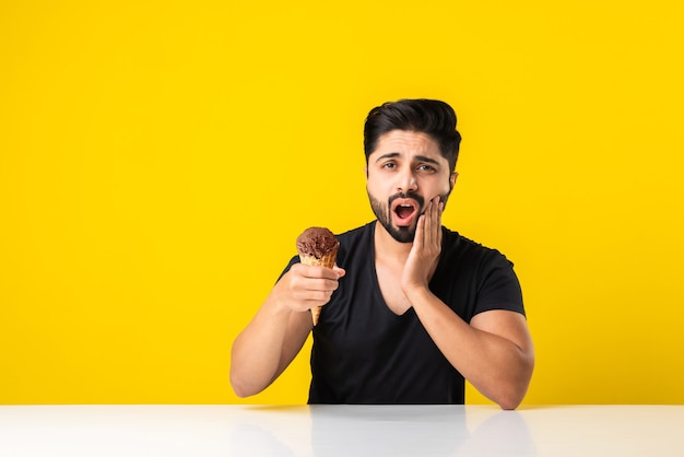 Giovane indiano che mangia il gelato nel cono e ha mal di denti a causa della cavità, seduto a tavola su sfondo giallo o bianco