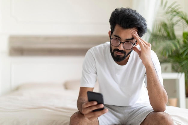 Giovane indiano stressato seduto sul letto e guardando lo schermo dello smartphone