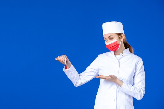 Giovane infermiera di vista frontale in vestito medico bianco con maschera rossa e iniezione nelle sue mani sulla parete blu
