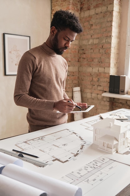 Giovane ingegnere architettonico afroamericano serio in maglione marrone in piedi al tavolo con un progetto e che esamina il piano del progetto di design sul tablet