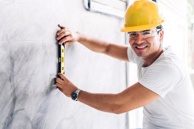 Giovane ingegnere di costruzione in un casco giallo che lavora e che effettua le misure sulla parete al cantiere della costruzione