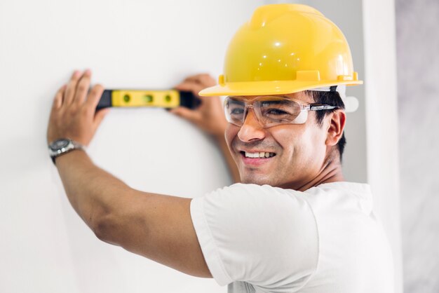 Giovane ingegnere di costruzione in un casco giallo che lavora e che effettua le misure sulla parete al cantiere della costruzione