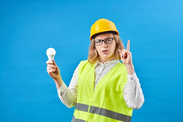 Giovane ingegnere femminile che indossa un giubbotto da lavoro e un casco giallo sulla testa che tiene la lampadina e punta il dito verso l'alto mentre è in piedi in studio su sfondo blu Il concetto di riparazione della costruzione