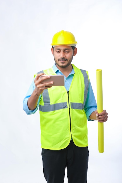 Giovane ingegnere indiano che indossa l'elmetto protettivo e utilizza lo smartphone su sfondo bianco.