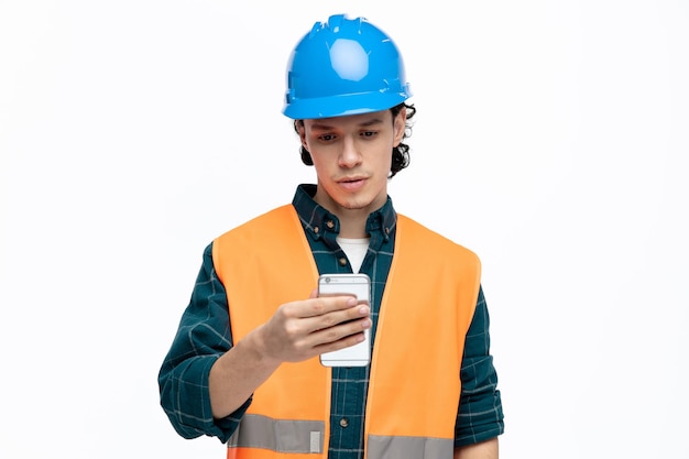 Giovane ingegnere maschio concentrato che indossa casco di sicurezza e giubbotto di sicurezza che tiene e guarda il telefono cellulare isolato su sfondo bianco