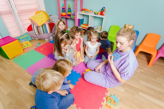 Giovane insegnante attraente che aiuta i bambini piccoli a essere coinvolti nelle loro prime lezioni alla scuola materna Educatore entusiasta con bambini piccoli su un pavimento mentre la matematica