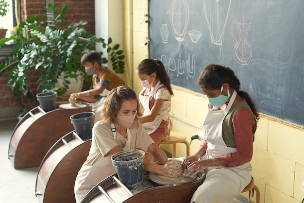 Giovane insegnante di ceramica in maschera che aiuta una ragazza nera a modellare l'argilla sul tornio in classe