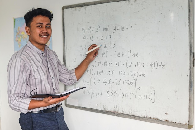 Giovane insegnante di matematica asiatica che tiene in mano libri e spiega formule matematiche scritte su tavola bianca