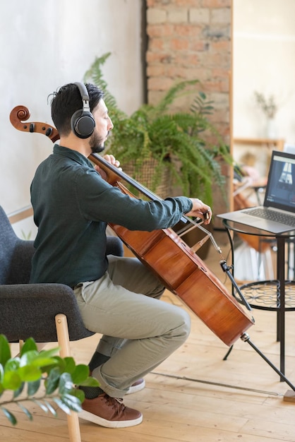 Giovane insegnante di musica che suona il violoncello davanti al computer portatile