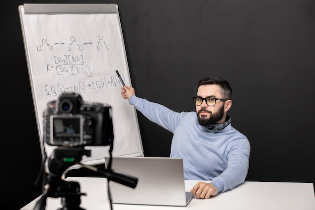 Giovane insegnante fiducioso che indica alla lavagna con formule chimiche durante la lezione online