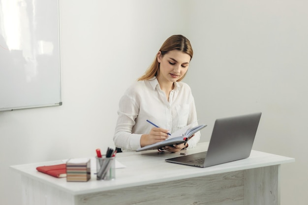 Giovane insegnante lavora al computer Indossa una camicia bianca Conduce corsi di formazione online per gli studenti Tiene in mano un taccuino Lezioni online Webinar