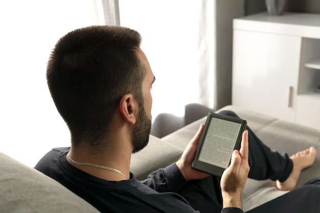 Giovane irriconoscibile che legge con il suo ebook mentre è seduto sul divano di casa
