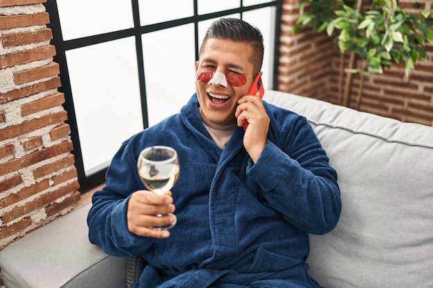 Giovane ispanico che indossa accappatoio e cerotti per borse sotto gli occhi bevendo vino parlando al telefono ammiccando guardando la telecamera con un'espressione sexy faccia allegra e felice