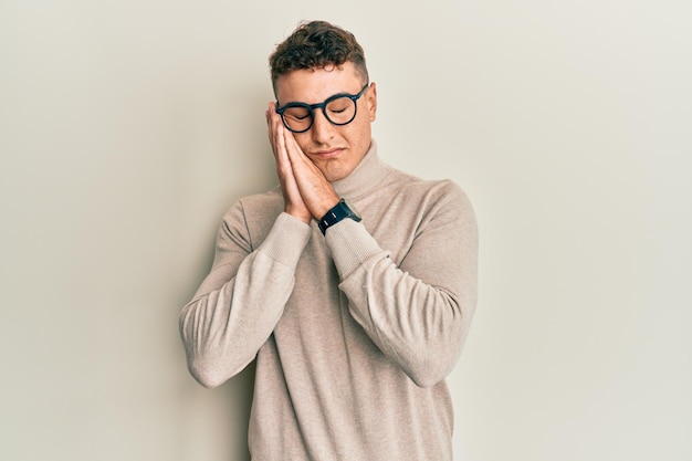 Giovane ispanico che indossa un maglione dolcevita casual che dorme stanco sognando e posando con le mani insieme mentre sorride con gli occhi chiusi