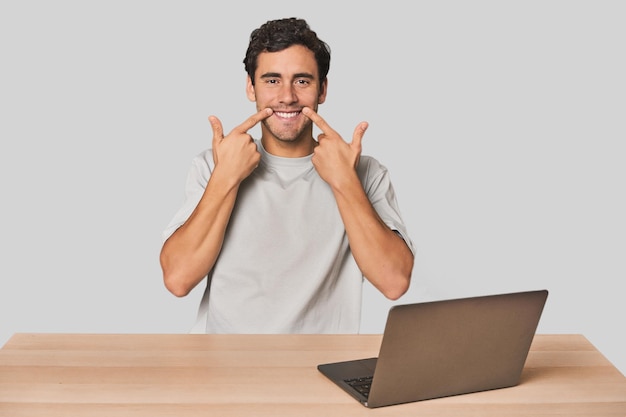 Giovane ispanico che lavora al portatile sorride indicando la bocca con le dita